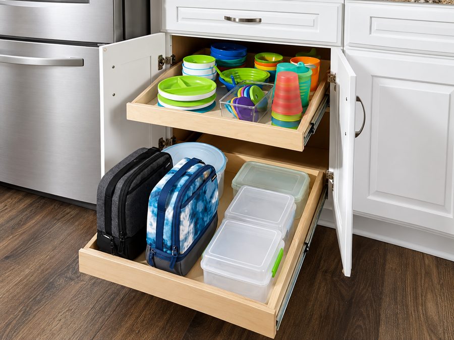 A base cabinet with both doors open and two maple pull out trays fully extended, holding stacked dishes and storage containers